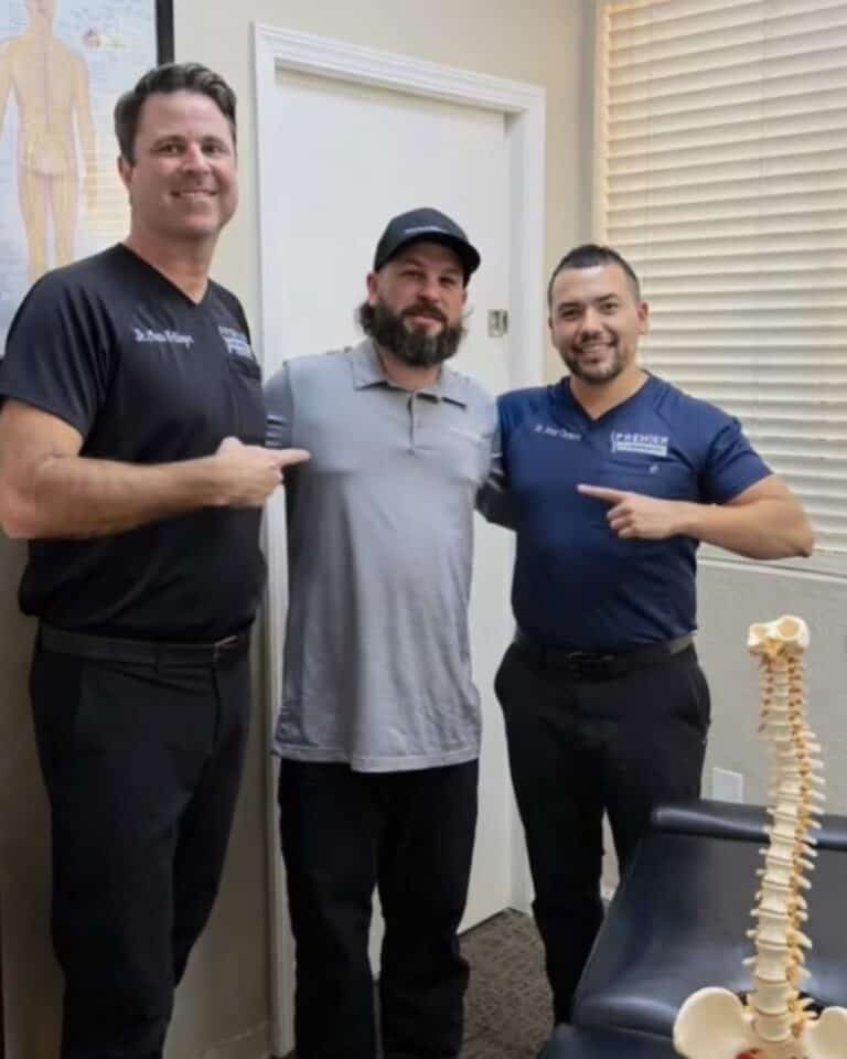 Dr. Christopher Schlenger and Dr. Jesús Cazares stand with a man in a gray polo shirt and black cap next to a spine model at Premier Chiropractic